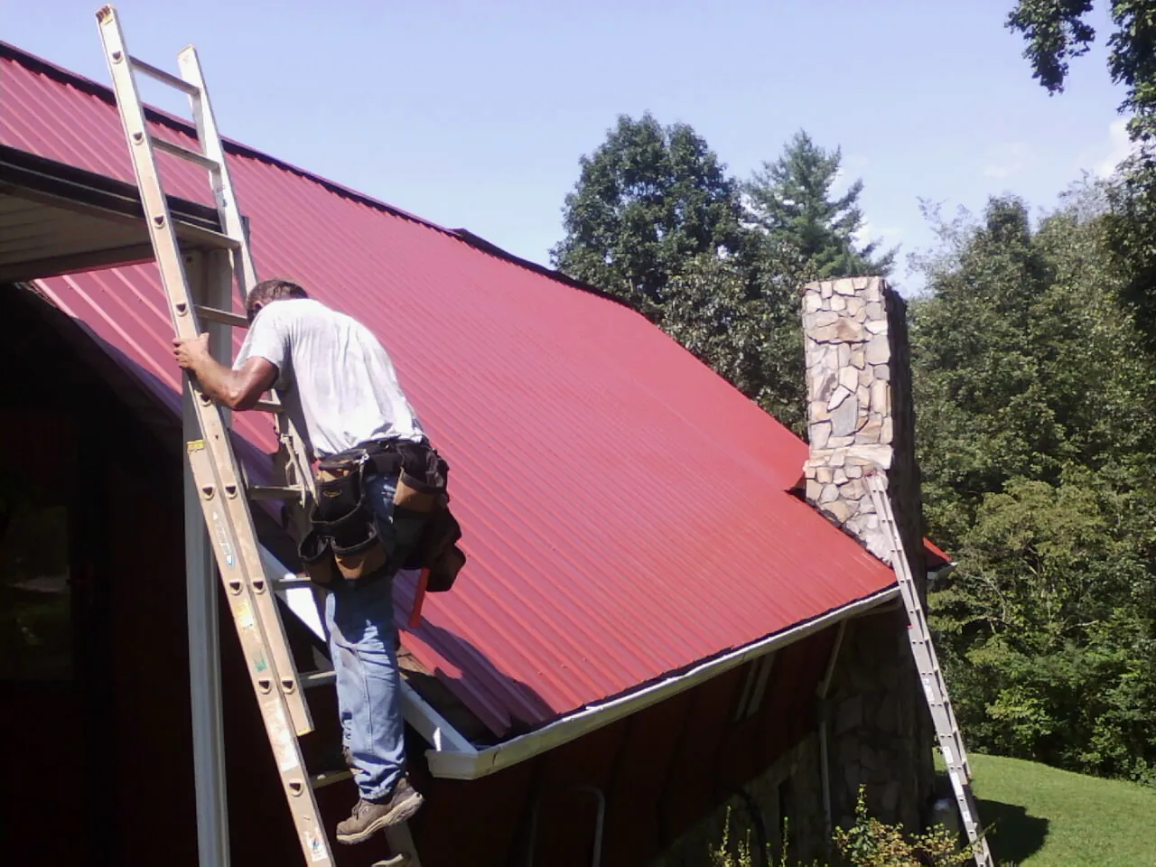 Roofer on a ladder beside a red metal roof and stone chimney — Soffit & Fascia Repair by Roofing Brothers in Oradell